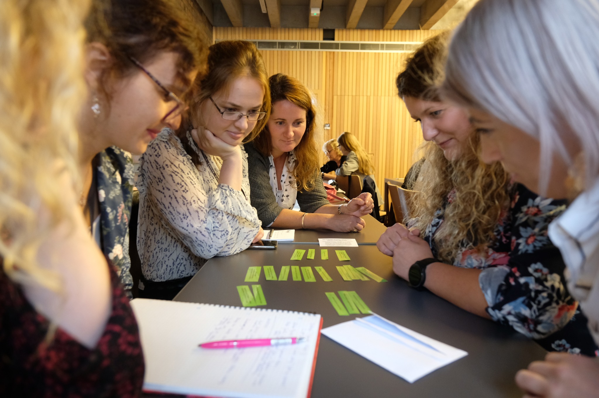 4 kilka kobiet pochyla się nad kolorowymi kartkami rozłożonymi na stole między nimi