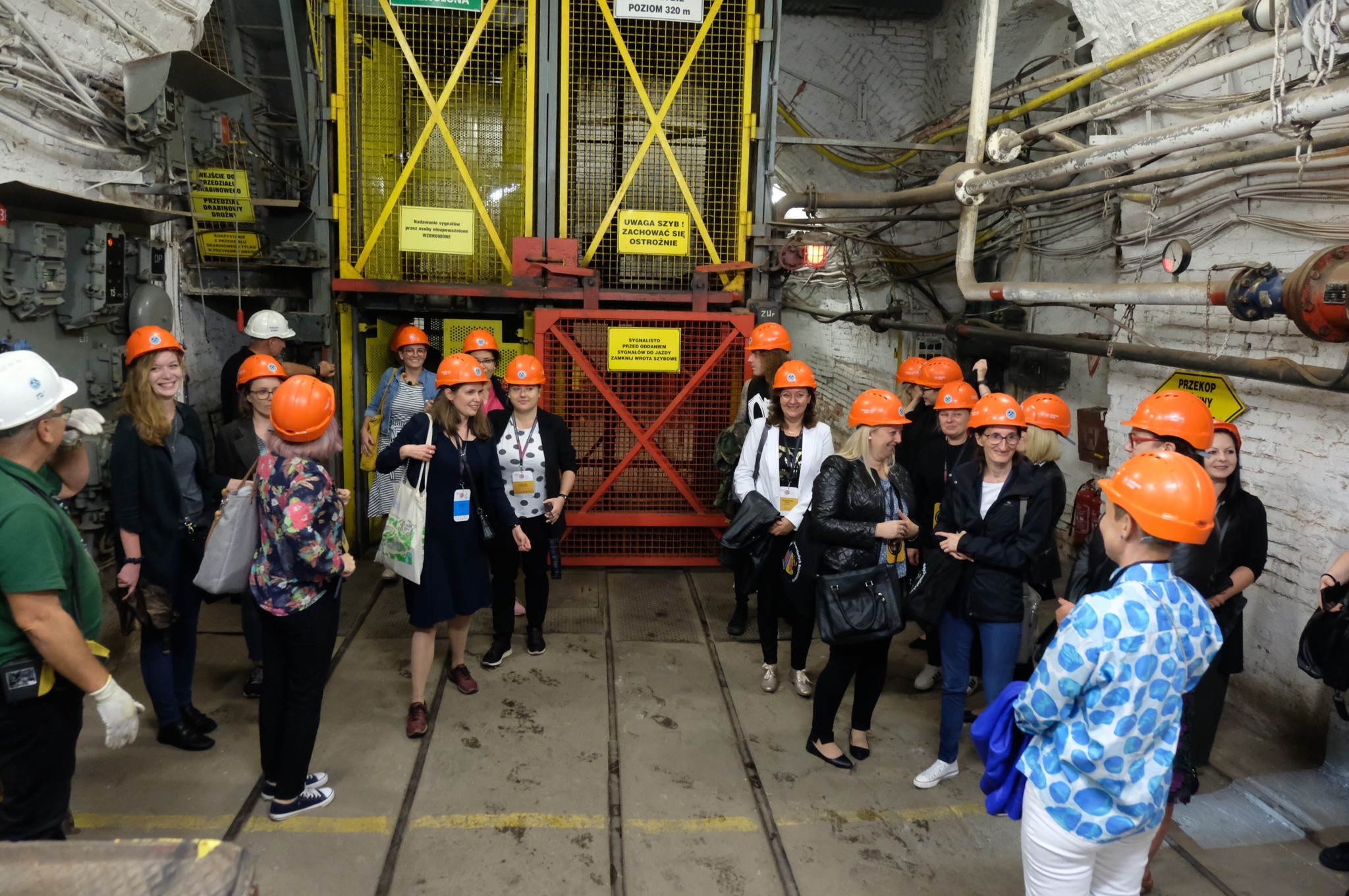 grupa bibliotekarzy w pomarańczowych kaskach stoi przy windzie w kopalni