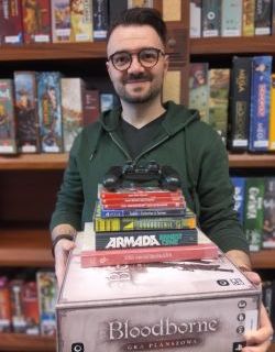 Sebastain Kochaniec, młody uśmiechnięty mężczyzna w okularach trzyma w ręku stos gier planszowych