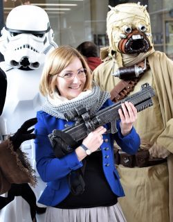 Karolina Jaskierska, młoda blondynka w okularach trzyma atrapę broni, za nią dwie postaci z Gwiezdnych Wojen