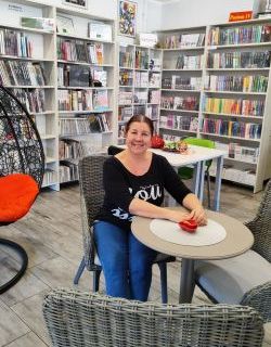 Anna Przygódzka, uśmiechnięta kobieta ze związanymi włosami siedzi przy stoliku otoczona regałami z książkami