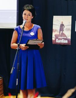 Agnieszka Opala, brunetka ubrana w kobaltową wizytową sukienkę stoi na scenie i mówi do mikrofonu