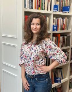 Młoda kobieta, Ludmiła Guzowska, w kręconych brązowych włosach i kwiecistej bluzce stoi przy regale z książkami