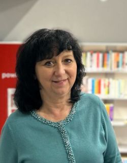Marzena Zeglin, Uśmiechnięta brunetka w długich włosach i błękitnej bluzce, w tle nowoczesna zaaranżowana na biało przestrzeń biblioteczna