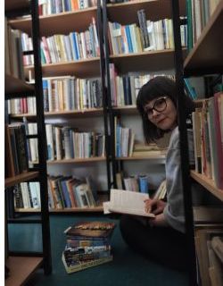 Ewa Michalak, młoda brunetka w okularach siedzi pod regałem trzymając otwartą książkę na kolanach