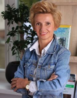 Agnieszka Kaczmarek, uśmiechnięta zdecydowana kobieta z krókimi zadziornie zaczesanymi włosami do góry, ma ręce skrzyżowane, w tle regał do ekspozycji 