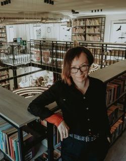 Kamilla Marchewska,  brunetka w okularach opiera się o poręcz bibliotecznej antresoli, wokół regały z książkami