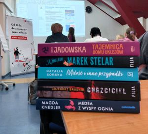 Grupa bibliotekarzy ogląda ksiazki ustawione na stołach, maj w rękach reklamowe torby, w le widać ekran prezentacji