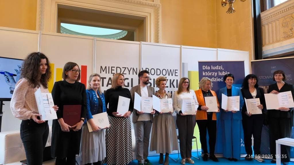 Grupa 11 bibliotekarek i bibliotekarzy trzyma dyplomy, stoi na scenie, w tle logo targów.