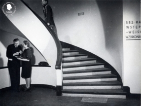 hol biblioteki, w górę prowadzą łukowate schody, które kontrastują z bielą ścian i białej poręczy. Obok schodów dyskutuje kobieta i meżczyzna.