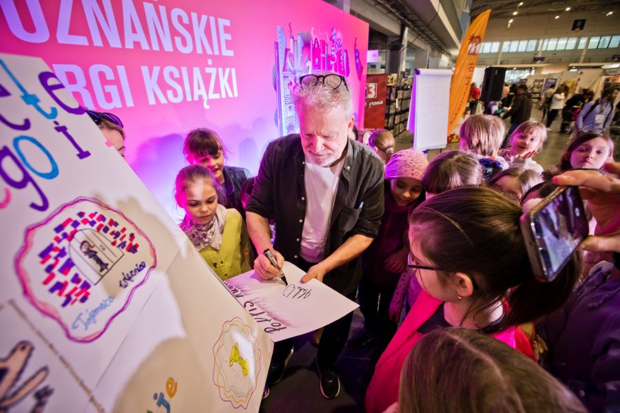 Pisarz Martin Widmark podpisuje plakat, otoczony przez grupę dzieci na Targach Książki w Poznaniu. W tle pomarańczowe stoisko z napisem 'Poznańskie Targi Książki