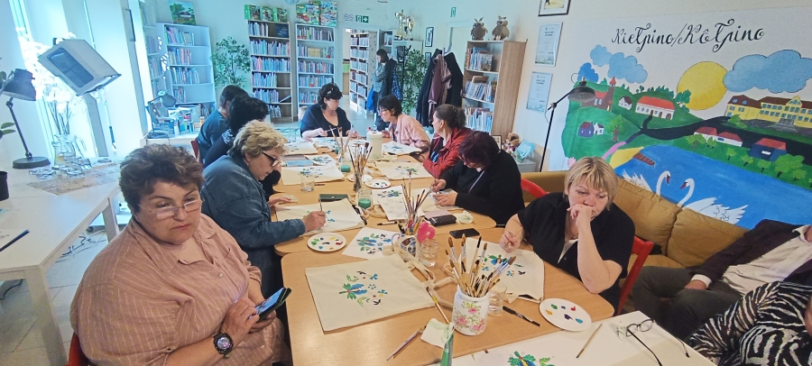 Grupa bibliotekarzy siedzi przy stole. Malują ręcznie torby.