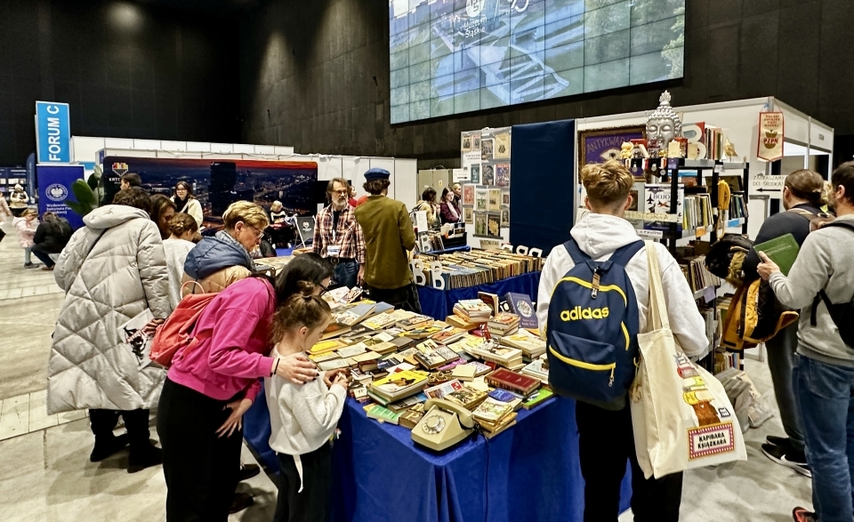 Stoiska targowe obłożone książkami. Międyz nimi ludzie pochylają się i wertują książki.