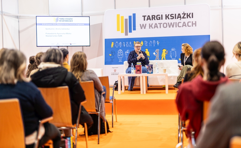 Spotkanie autorskie. Dwie kobiety rozmawiają na tle baeru z napisem Targi ksiązki w Katowicach.