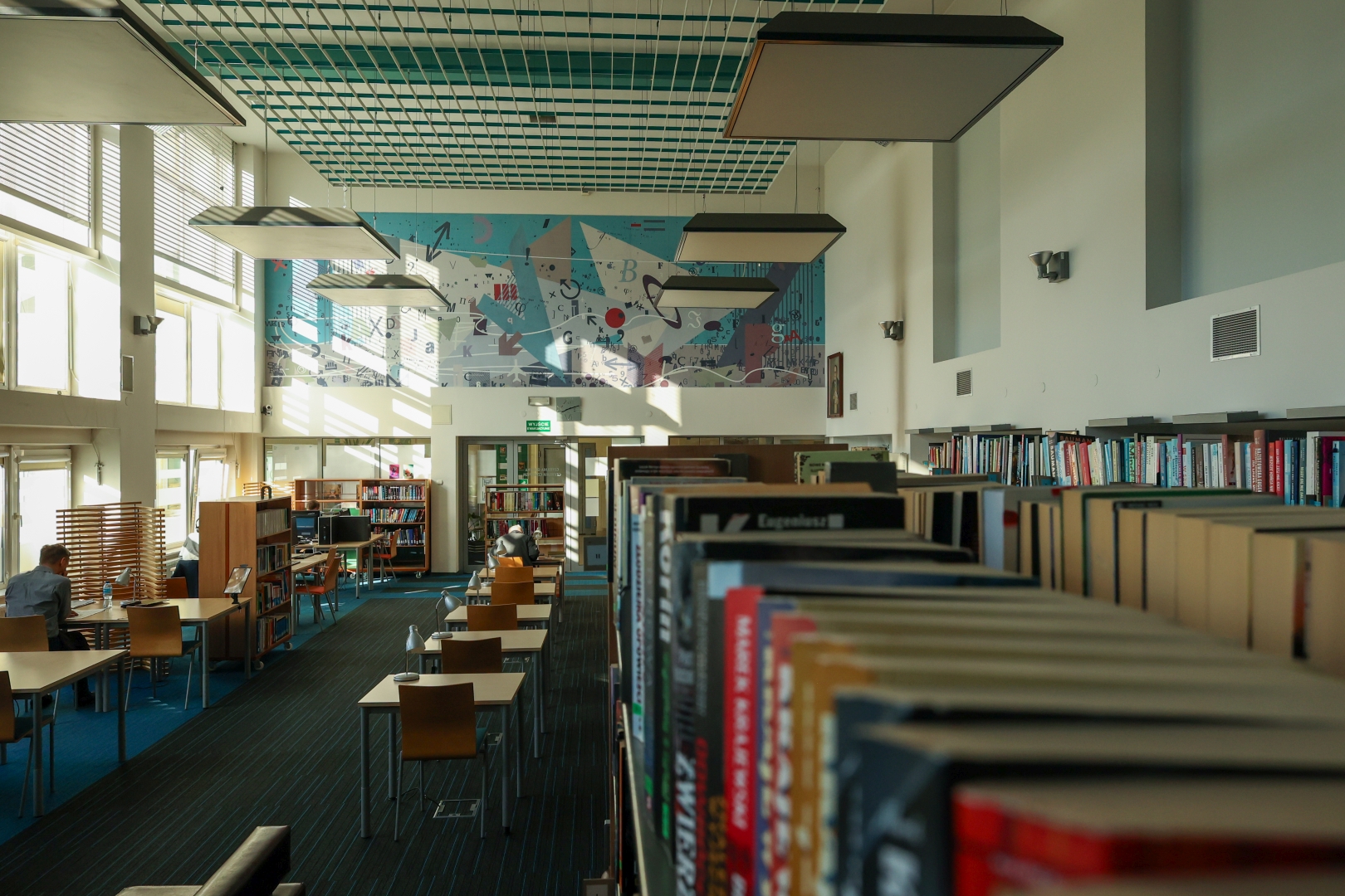 Wnętrze biblioteki. Sala z czytelni ze stolikami. Na ścianie szczytowej kolorowy mural. po prawejs stronie szereg regałów z książkami