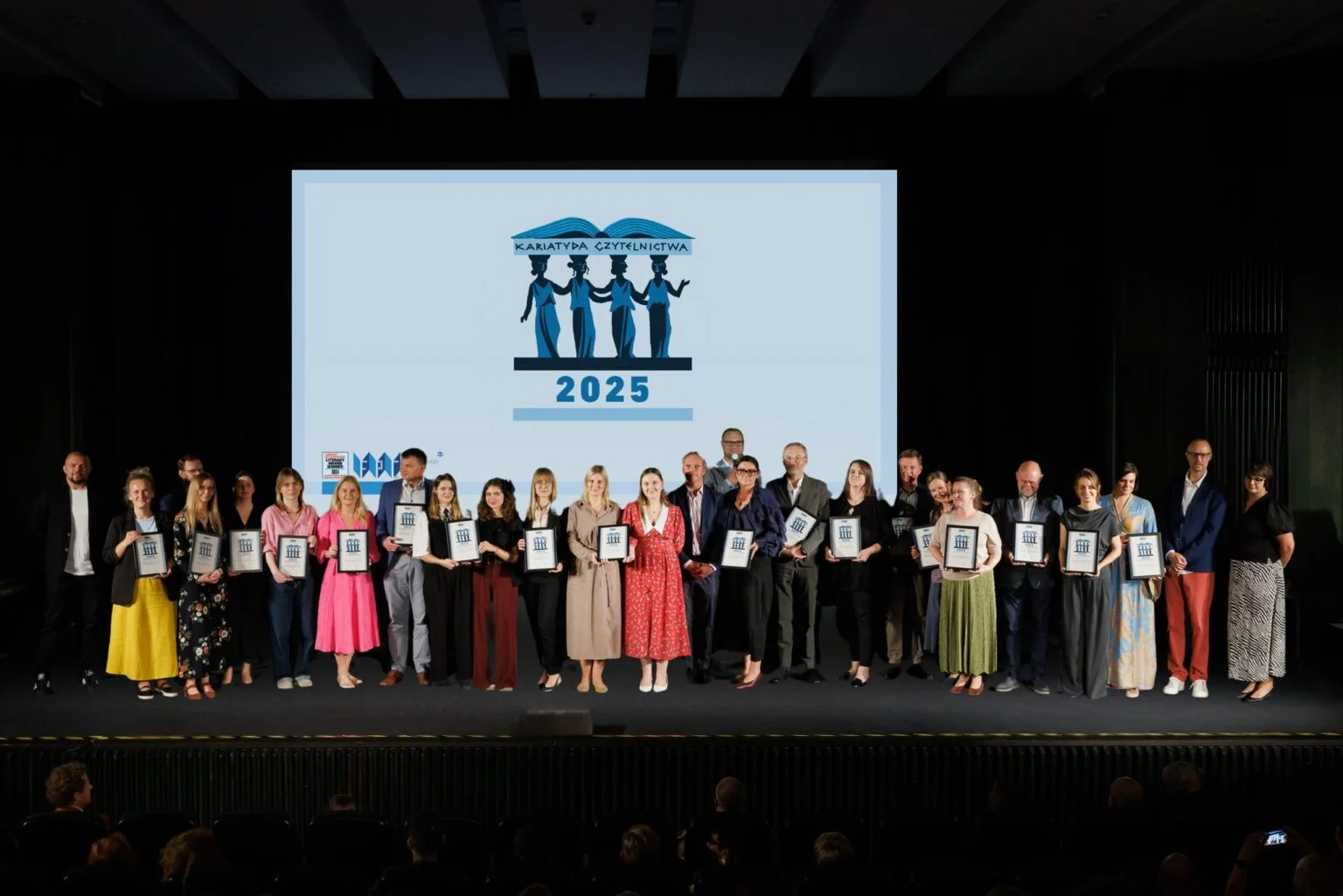 Grupa nagrodoznych osób stoi na scenie, trzyma dyplomy. W tle logo nagrody kariatydy czytelnictwa