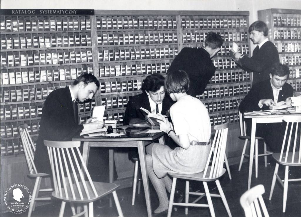 pomieszczenie z katalogami bibliotecznymi. Przy stoliku siędzą i notują dwaj mężczyźni i kobieta. Dwóch młodych mężczyzn sprawdza coś w katalogu.
