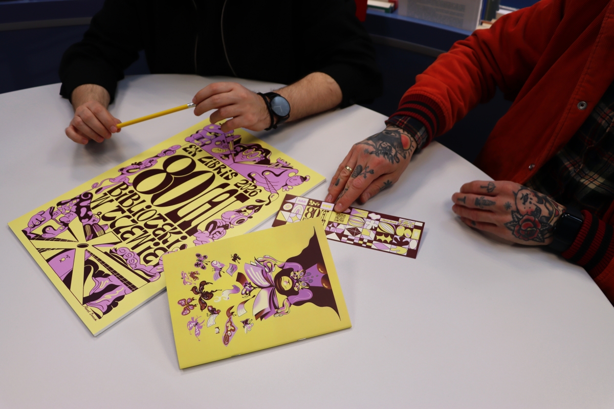Na stole leżą notes, grafika z ekslibrisem osiemdziesięciolecia biblioteki, ołówek. i zakładka do książki. Wszystko żółto-fioletowe.