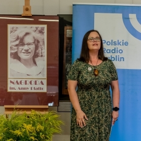 Brunetka w zielonej sukni i okularach stoi obok staerego zdjęcia bibliotekarki Anny Platto. pod zdjęciem podpis Nagroda im. Anny Platto.