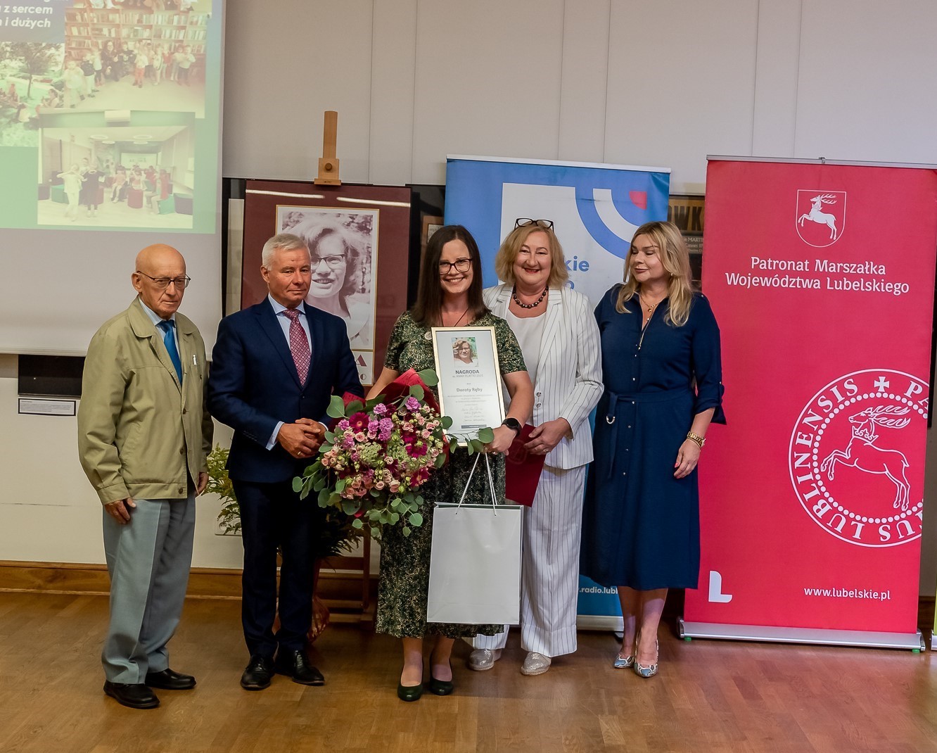 Laureatka nagrody trzyma dyplom i kwiaty. obok wręczający nagrodę.