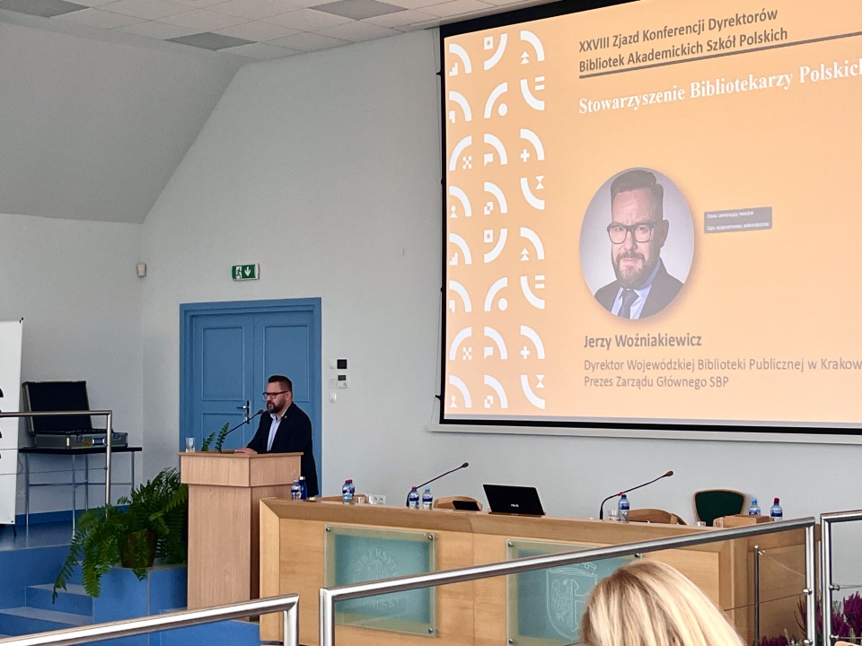 Sala wykładowa. Przewodniczący stoi przy mówinicy. W tele ekran z jego zdjęciem i afiliacją. 