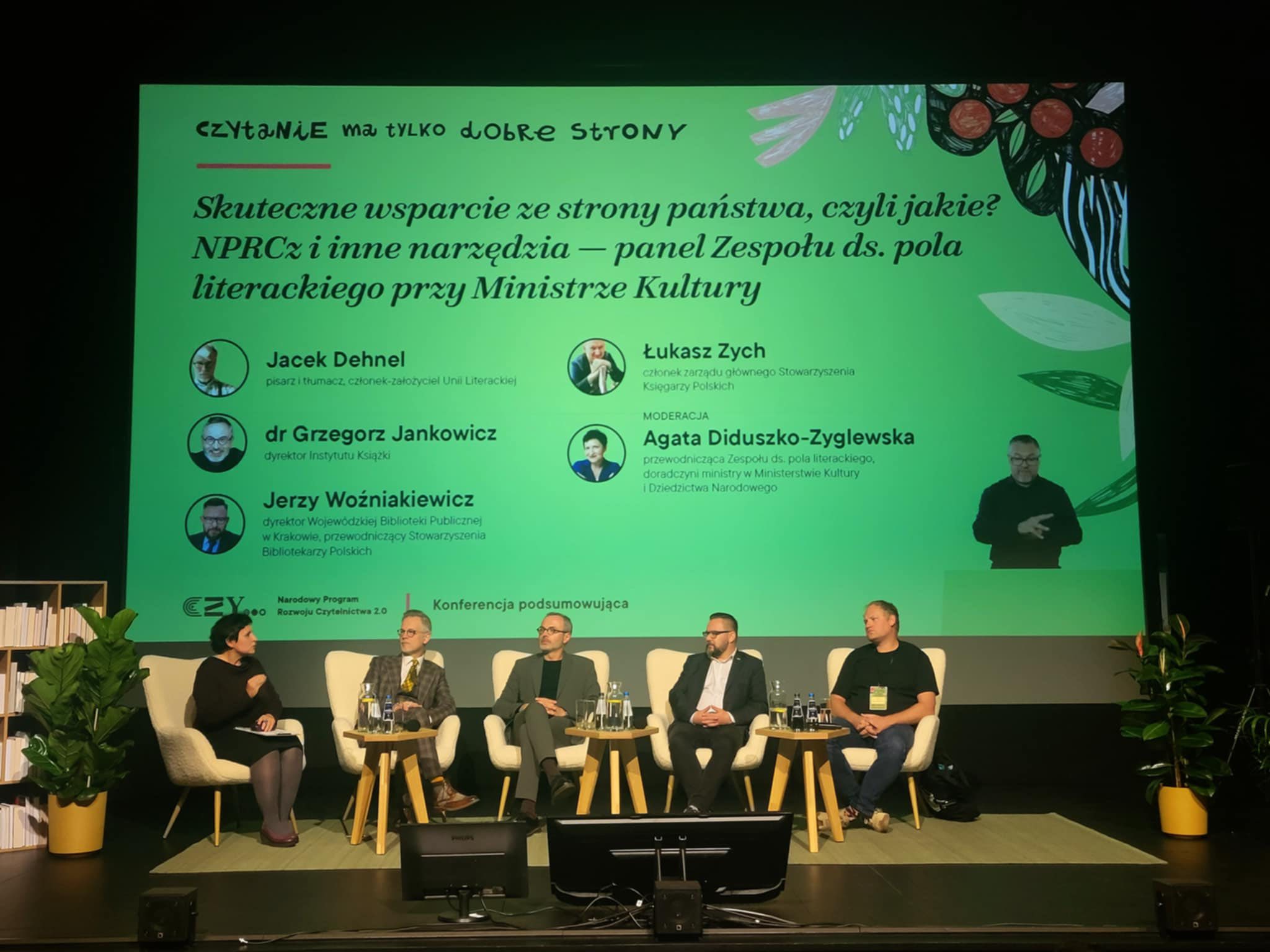 Czterech mężczyzn i kobieta siedzi podczas debaty na scenie. W tle ekran z tytułem panelu dyskusyjnego.