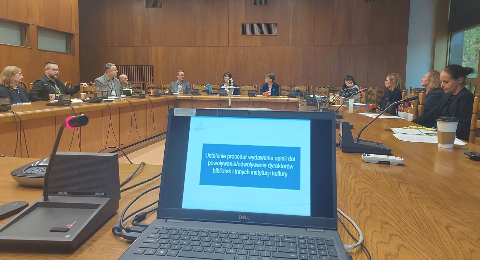 Sala konferencyjna, członkowie zarządu siedzą wokół stołu w literę U, na pierwszym planie komputer.