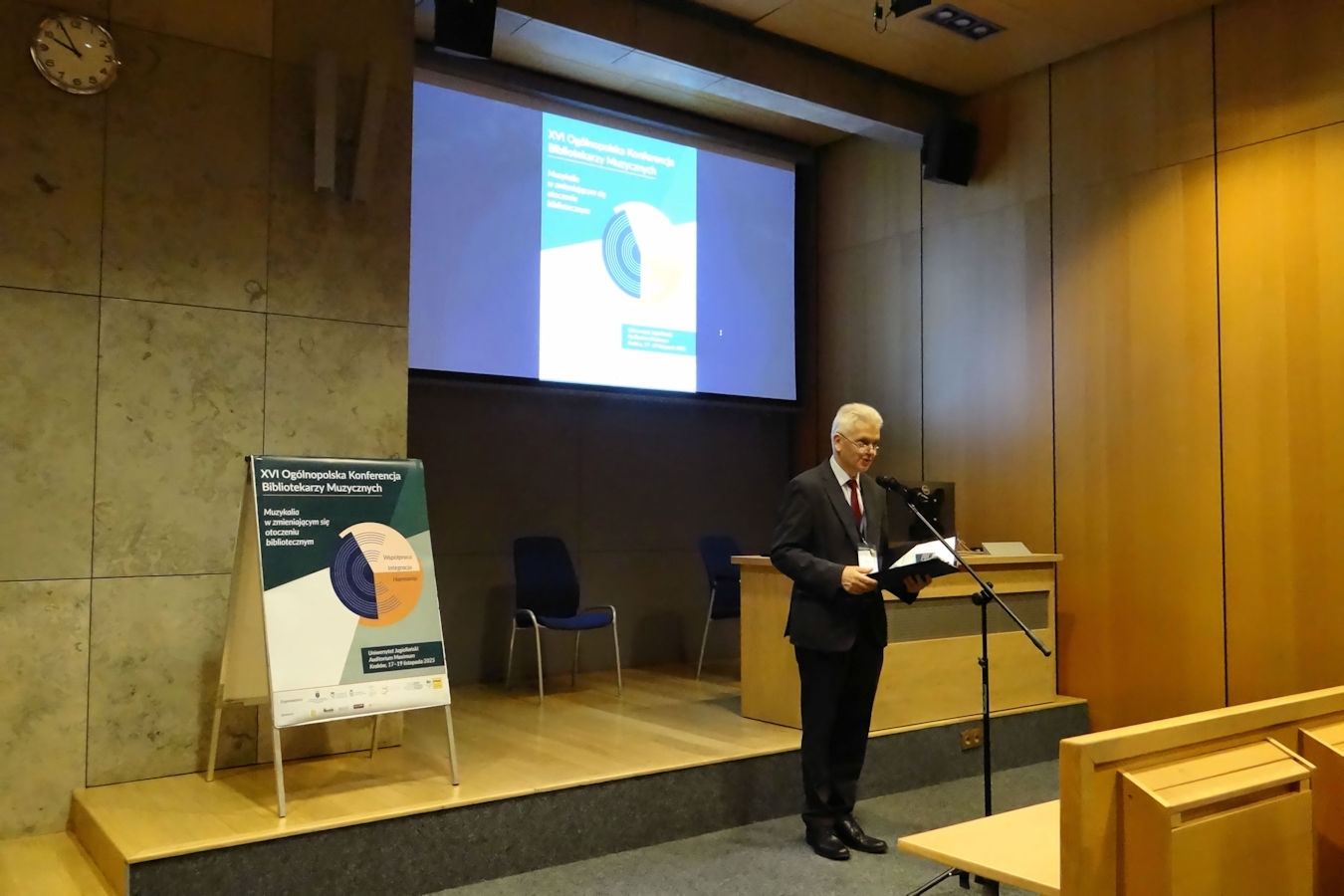 Mężczyzna stoi przed mikrofonem. Wita gości konferencji, w tle ekran